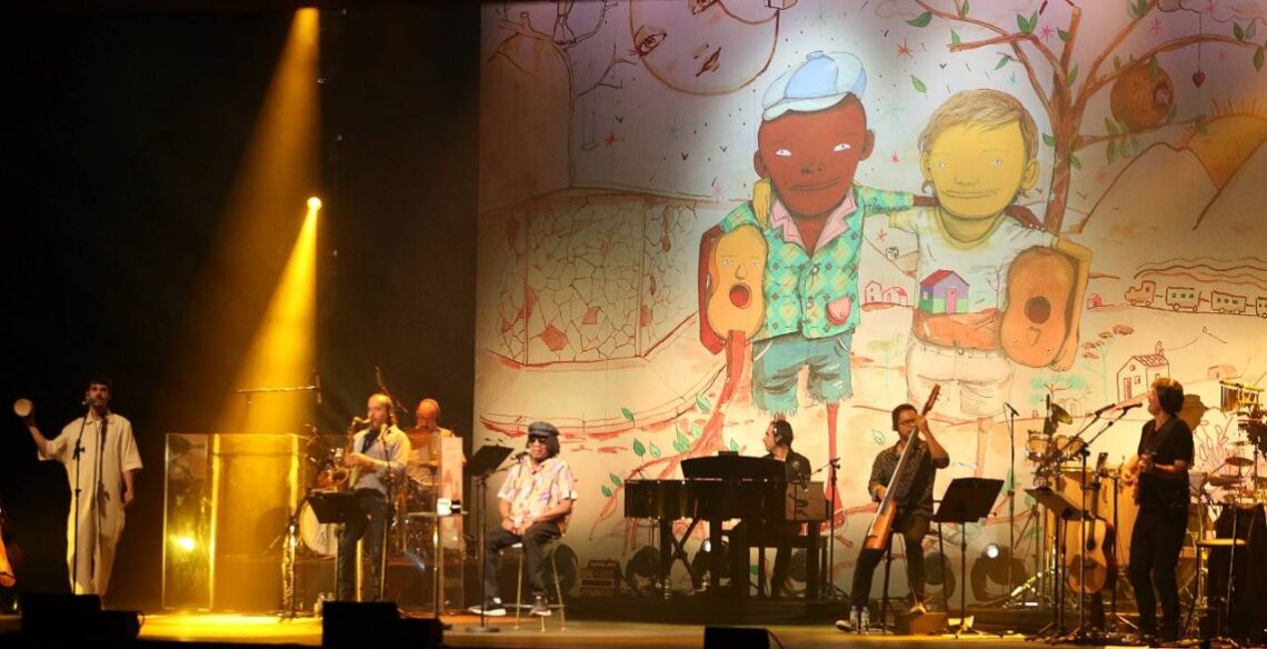 Cantor Milton Nascimento no palco com toda sua banda. Ao fundo uma imagem que faz referência ao album Clube da Esquina, um desenho dele com Lô Borges abraçados.