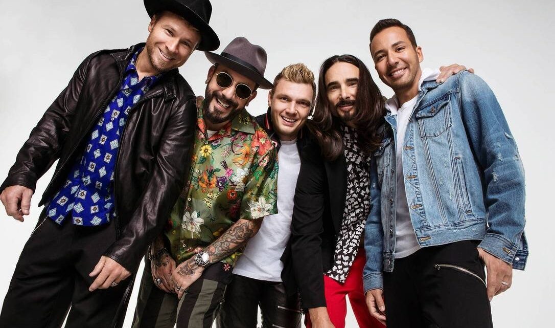 Banda Backstreet Boys posa para foto em fundo branco e com roupas coloridas, rindo e se abraçando
